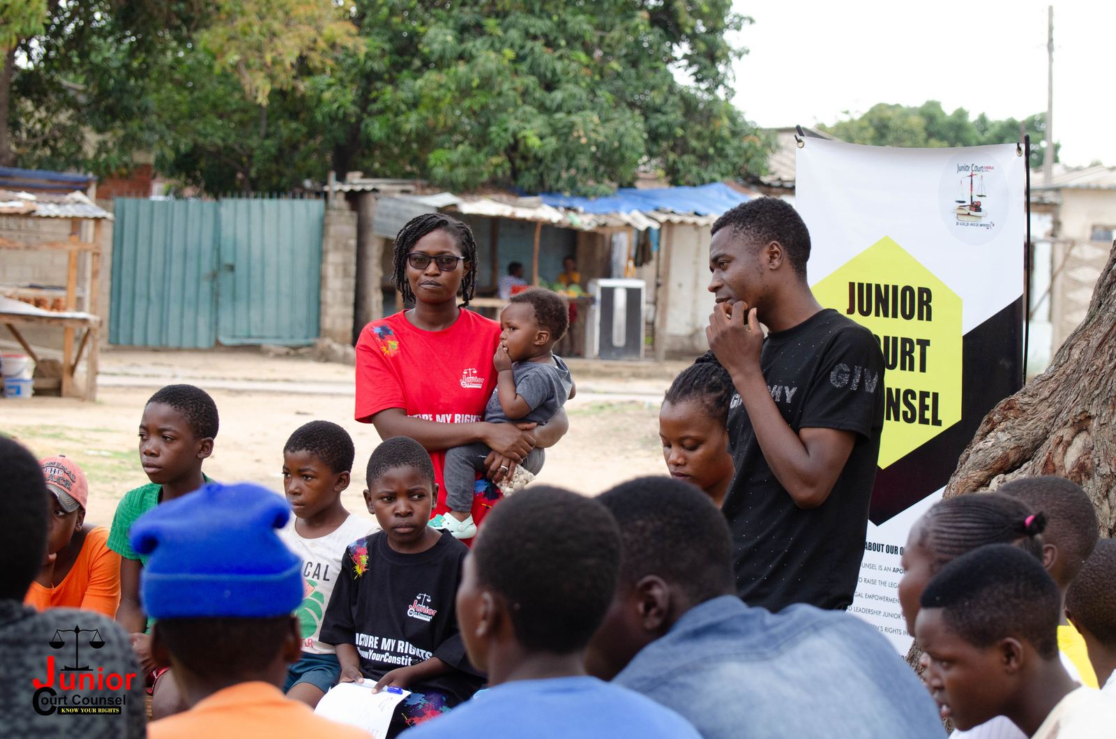 A volunteer holding a young child talks with a community member outdoors, with a Junior Court Counsel banner nearby