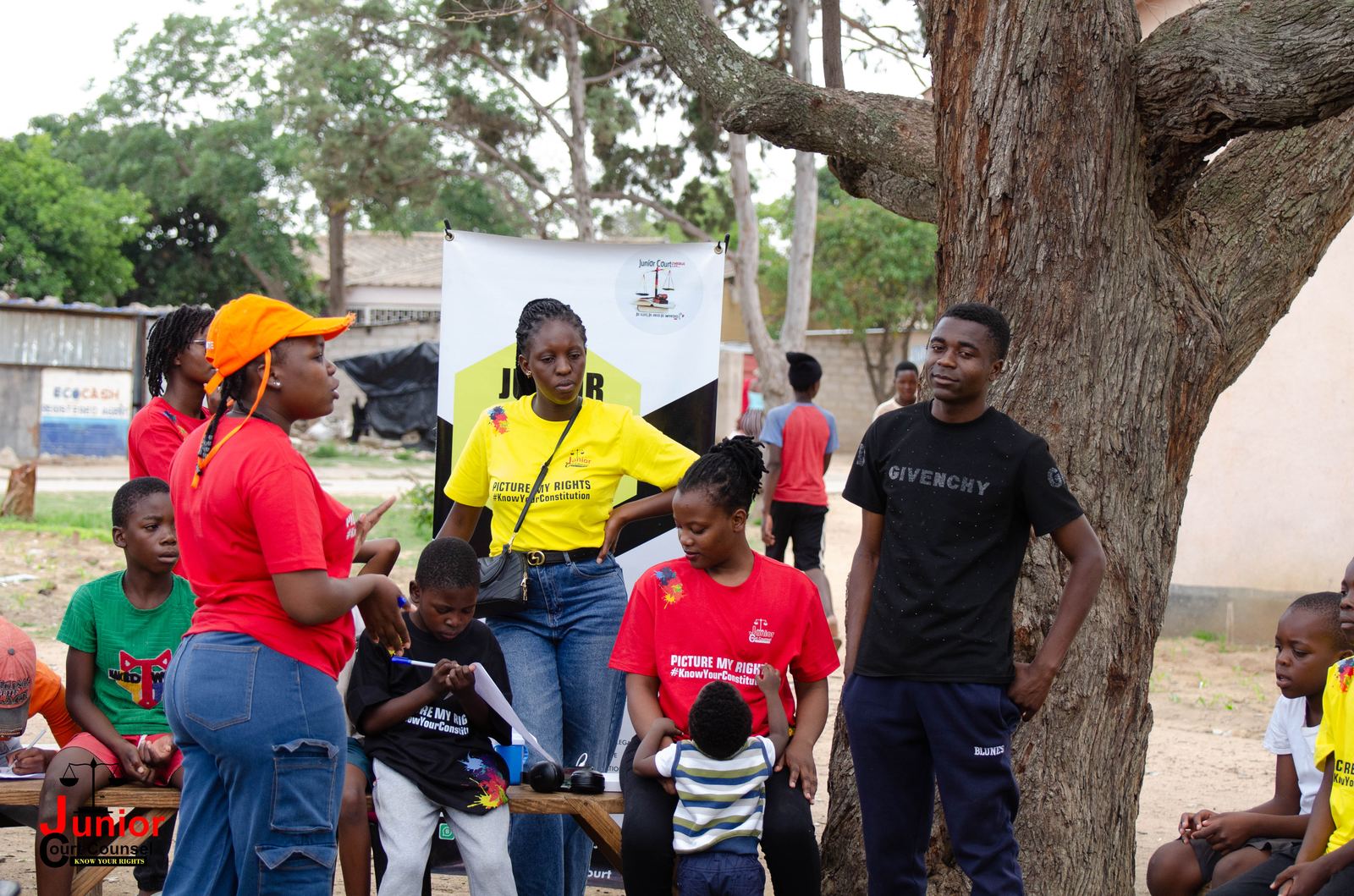 Four JCC volunteers in red and yellow t-shirts standing and talking under a tree beside campaign banners