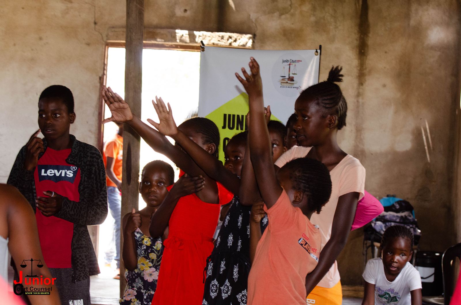 Children raising their hands to answer a question inside a community hall, with a Junior Court Counsel banner in the background