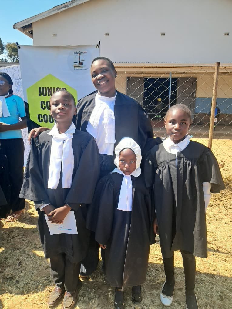 A JCC team member poses with three young participants in Junior Court robes beside the Junior Court Counsel banner