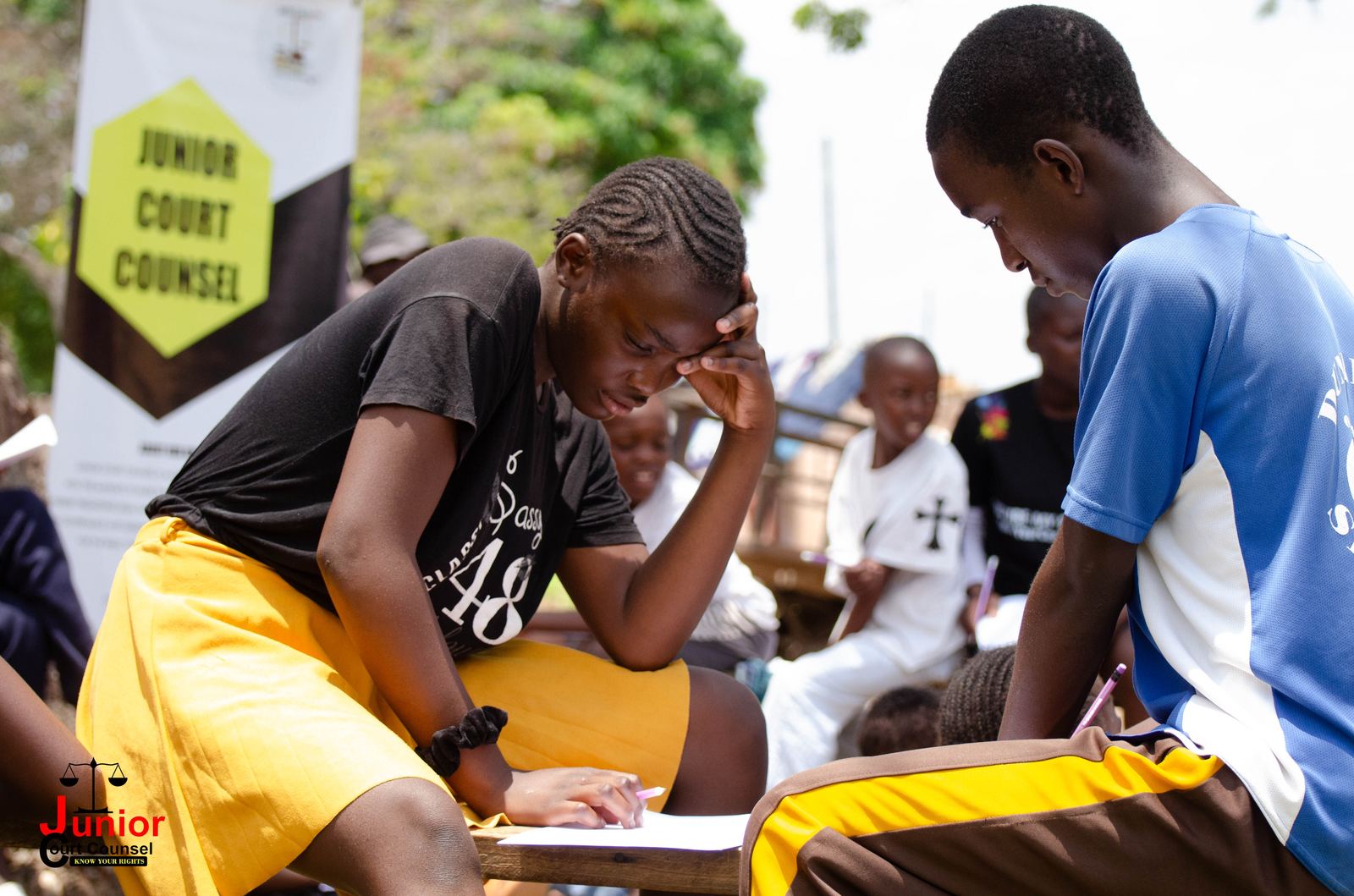 A teenager pausing to think while writing, with another participant leaning in beside a Junior Court Counsel banner