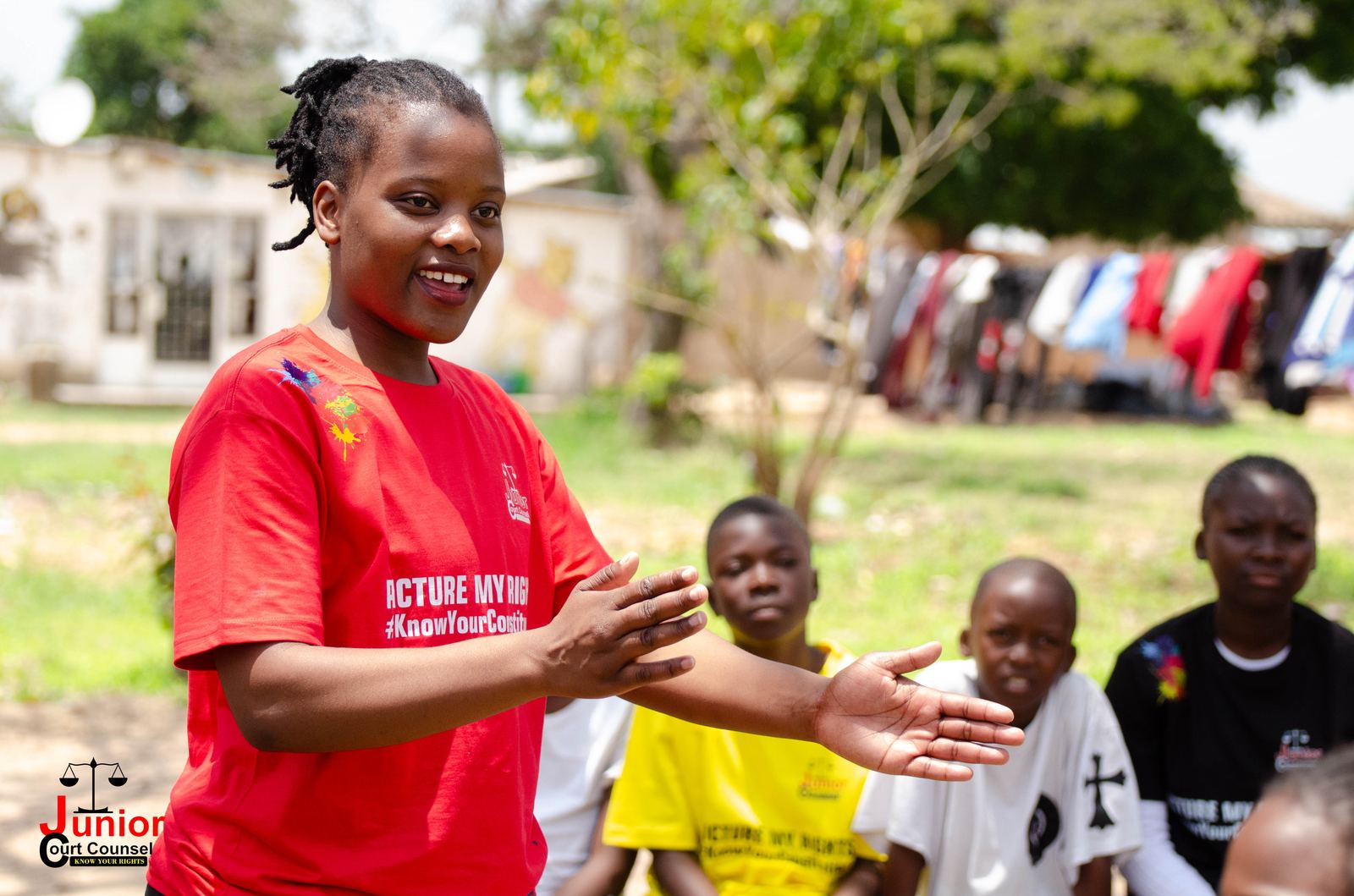 A JCC volunteer in a red campaign t-shirt gesturing while speaking to a group of children outdoors